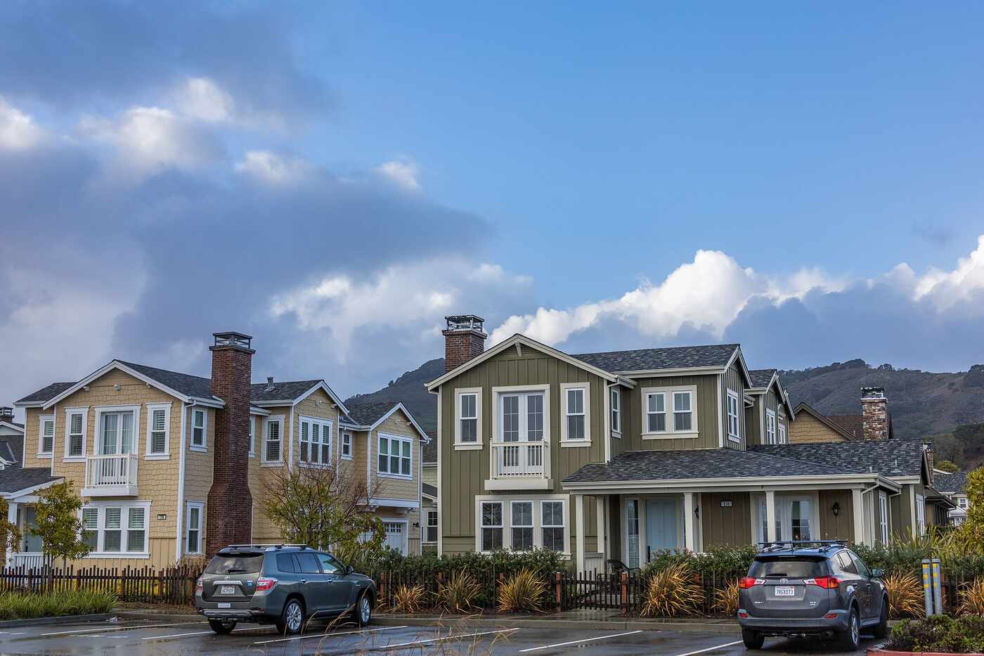 A residential street of settled houses with parked cars under a partly clouded sky
