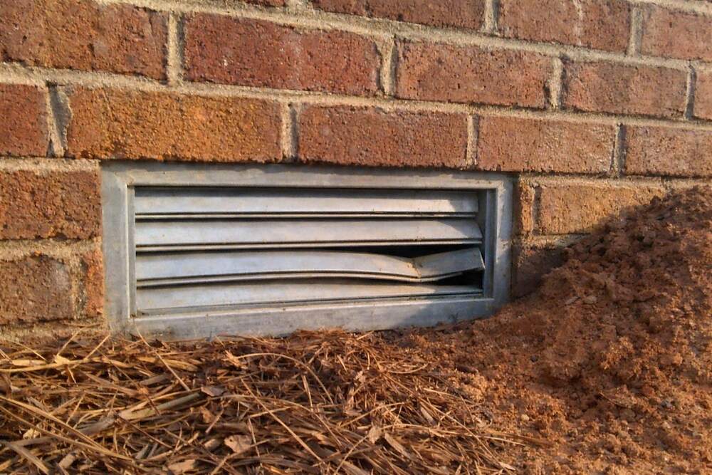 A crawl space vent set low in a brick wall with mulch and clay banked against it