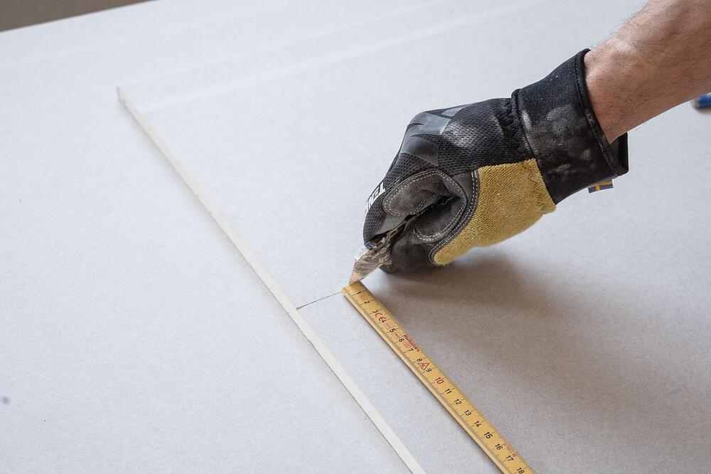 A gloved hand marking a straight line across a drywall sheet with a ruler